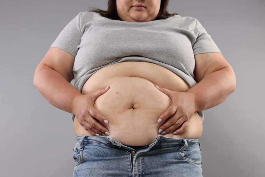 A person wearing a gray shirt and unbuttoned jeans holds the soft folds of their abdomen against a plain gray background. The close view represents bloating and concern about overnight weight gain.