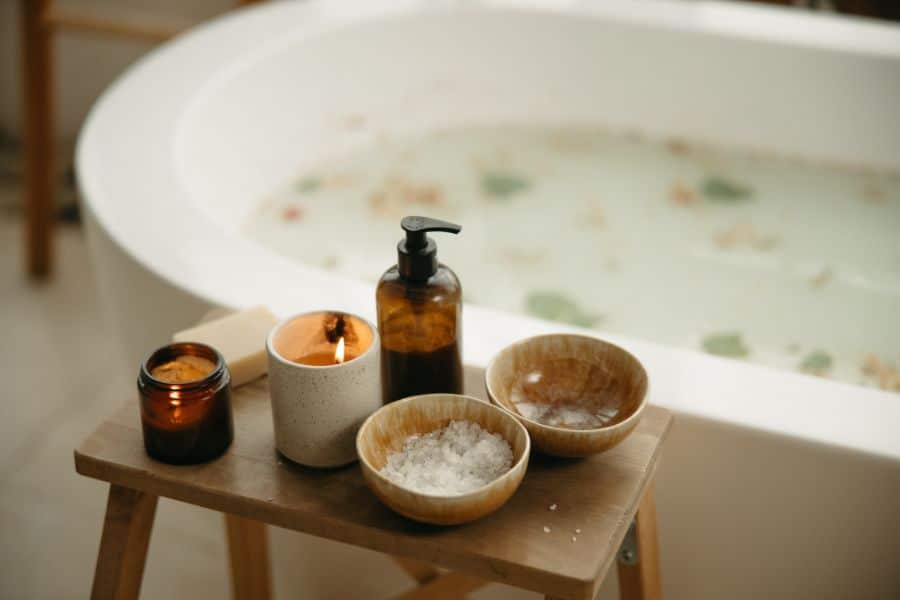 A warm bath is prepared with lit candles and bowls of bath salts on a wooden stool beside the tub. The calming setup represents a relaxing evening routine that can support better sleep after 50.