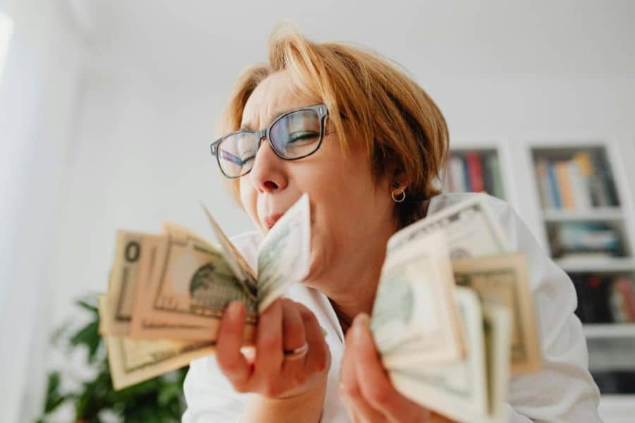 A middle aged womam with short hair and glasses holding two fists full of dollar notes