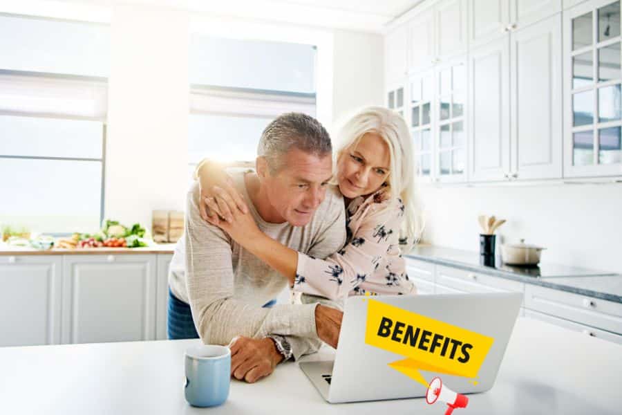 An older couple looks at a laptop together in a bright kitchen. The laptop displays the word "BENEFITS" with a megaphone graphic.
