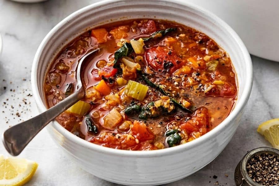 Hearty tomato based soup filled with lentils carrots celery leafy greens and other vegetables. Lemon wedges and cracked black pepper accompany the colorful vegetable soup.