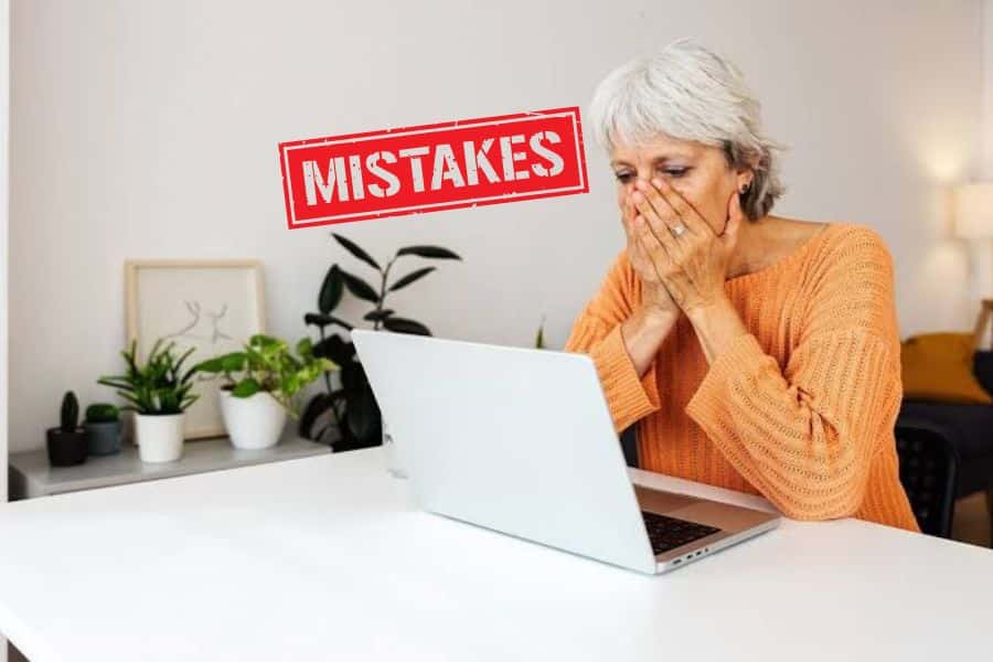 A woman with grey hair and wearing an orange top sat in front of a laptop with her hands over her mouth