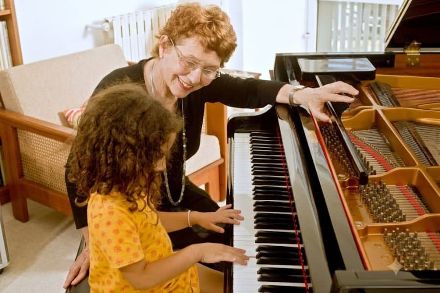 An older woman smiles while guiding a young child at a grand piano. Teaching piano offers a meaningful retirement hobby that combines music with time spent helping others learn.