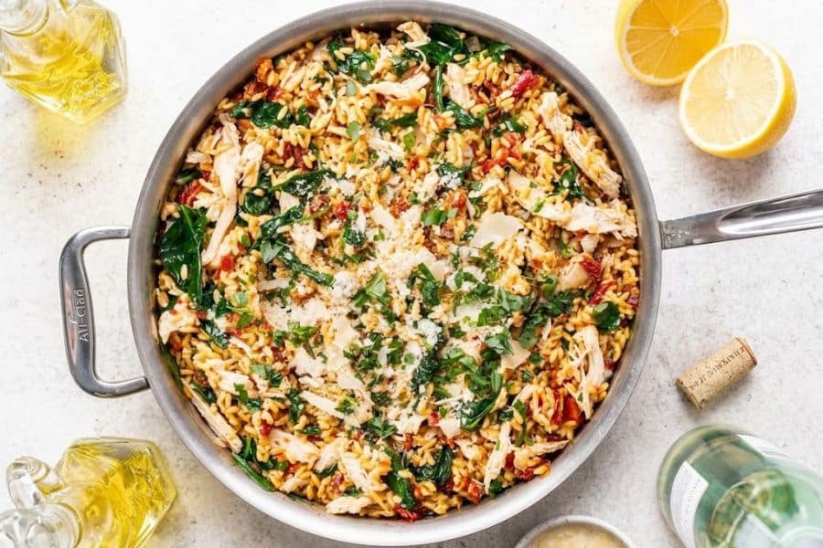 One pan chicken and rice with leafy greens tomatoes fresh herbs and grated cheese. Lemon and olive oil surround the skillet for a bright Mediterranean style meal.