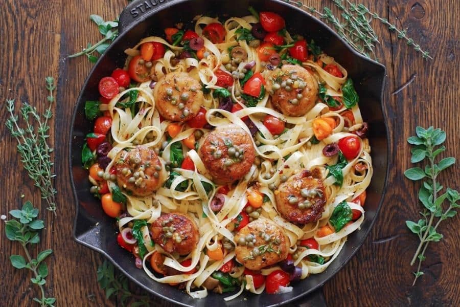 Wide noodles tossed with seared scallops cherry tomatoes spinach olives capers and fresh herbs in a cast iron skillet. The seafood pasta combines vegetables and seafood associated with Mediterranean diet cooking.