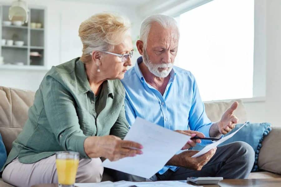 Older couple sitting together on a sofa while reviewing financial paperwork and discussing the documents in front of them. The scene supports a retirement withdrawal order discussion by showing retirees working through financial decisions together.