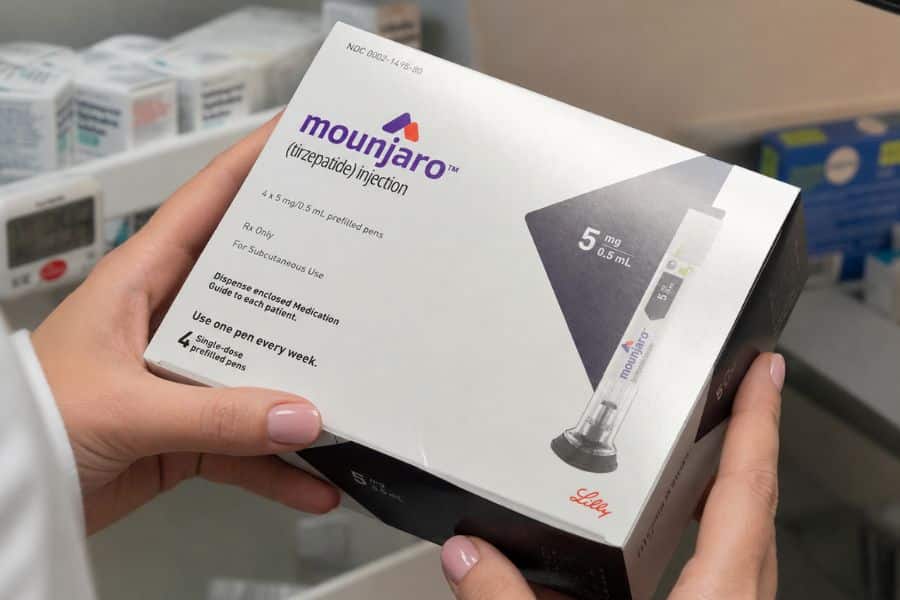 A box of Mounjaro tirzepatide injection is held inside a pharmacy during a discussion of weight loss medication. The packaging reads "NDC 0002 1433 80. Mounjaro. Tirzepatide injection. 4 by 5 mg per 0.5 mL prefilled pens. Rx Only. For Subcutaneous Use. Dispense enclosed Medication Guide to each patient. Use one pen every week. 4 single dose prefilled pens. 5 mg per 0.5 mL. Lilly."
