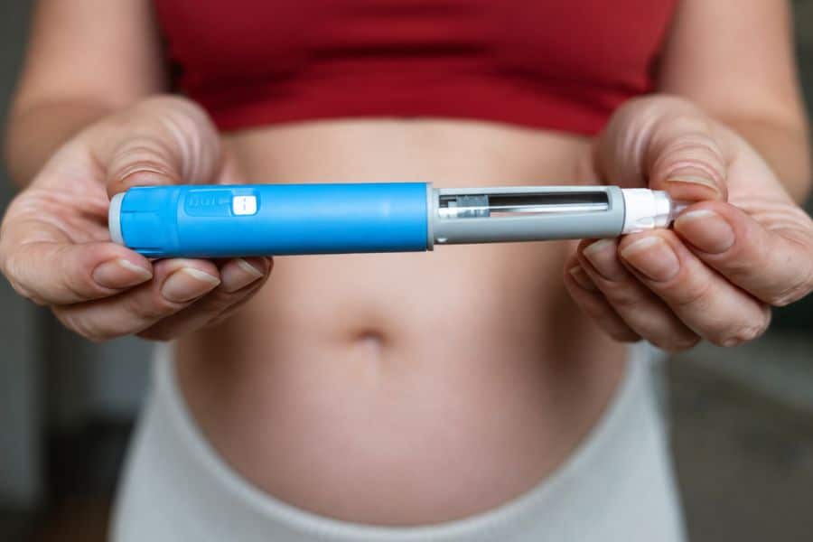 A person with a bare abdomen holds a blue and gray injectable medication pen for weight-loss jabs. The clear section reveals the medication chamber and internal mechanism.