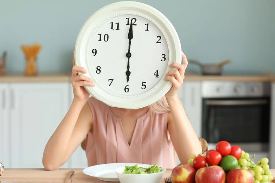 A woman sat at a kitchen table with breakfast in front of her and a big clock hiding her face