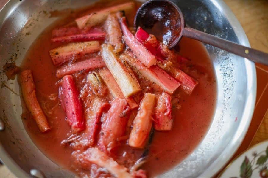 Chopped pink and red rhubarb stalks cook in a reddish syrup inside a metal pan. A dark wooden spoon rests across the softened rhubarb.
