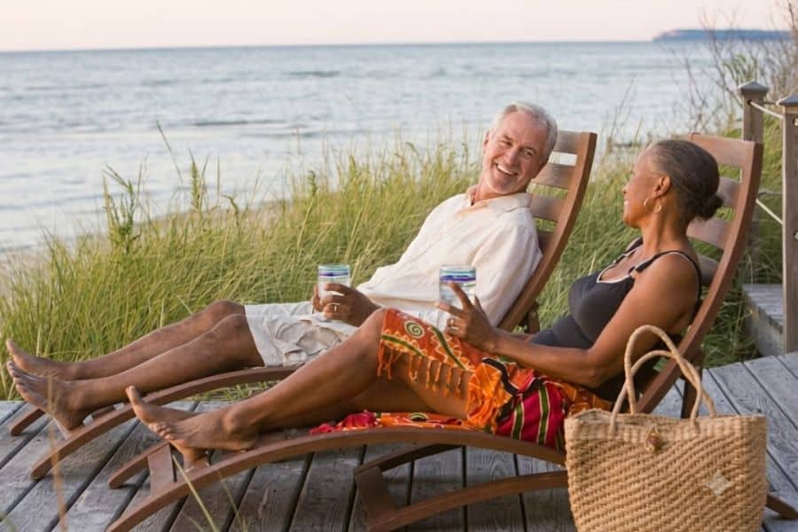 Retired couple relaxes in reclining chairs on a beachside deck while chatting and holding drinks with the ocean behind them. The peaceful setting shows enjoying leisure time and building a fulfilling lifestyle after the retirement slump.