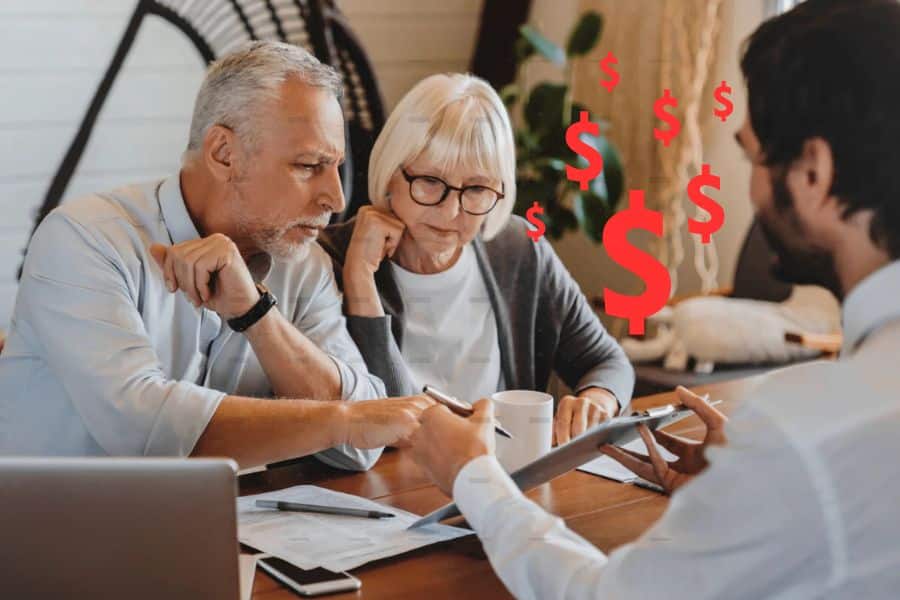 An older couple sits at a table reviewing financial documents with an advisor. Red dollar signs hover between them to emphasize a discussion about money and finances.