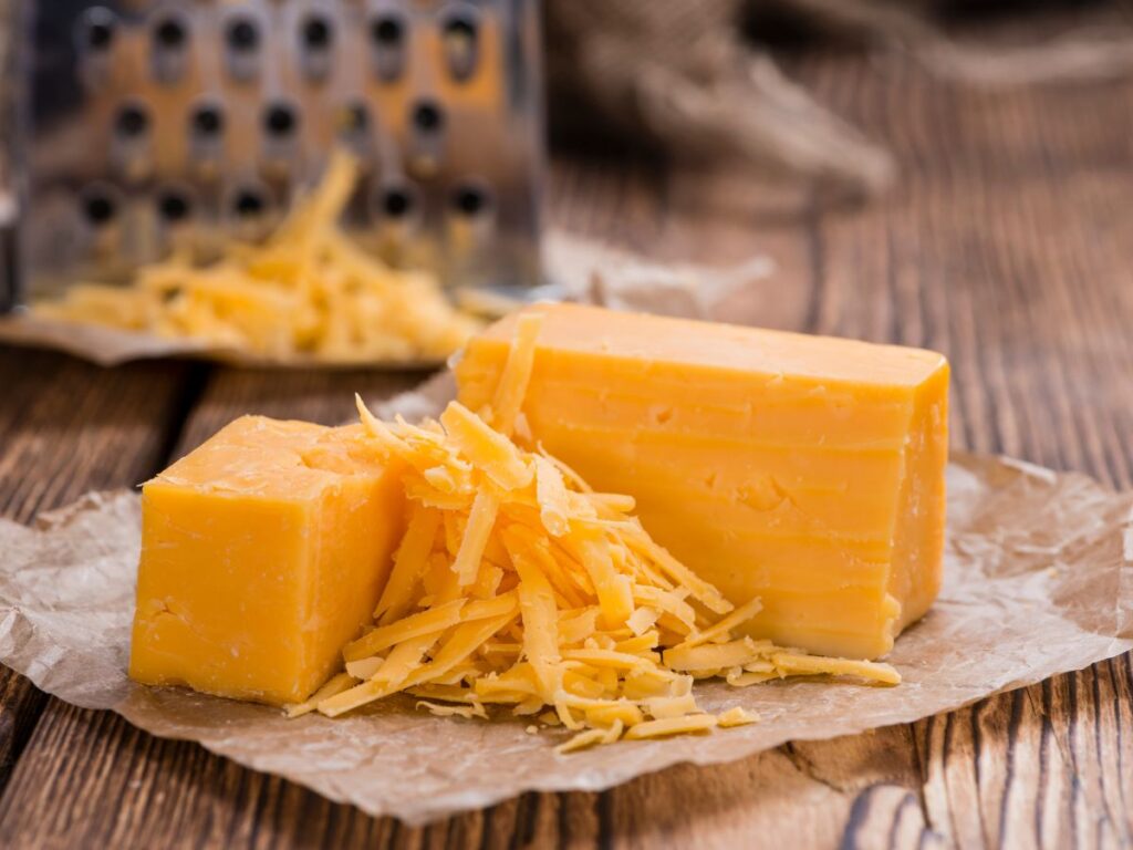 Blocks of orange cheddar cheese sit beside a pile of freshly shredded cheese on parchment paper with a box grater in the background. Cheddar provides a protein rich option for meals focused on foods that help rebuild muscle.