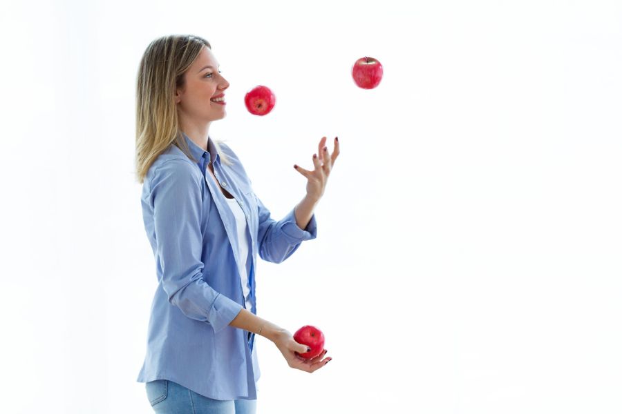 A smiling woman juggles three red apples against a plain white background. The playful coordination exercise suggests keeping the mind and body active as part of learning How To Raise Your IQ Over 50.