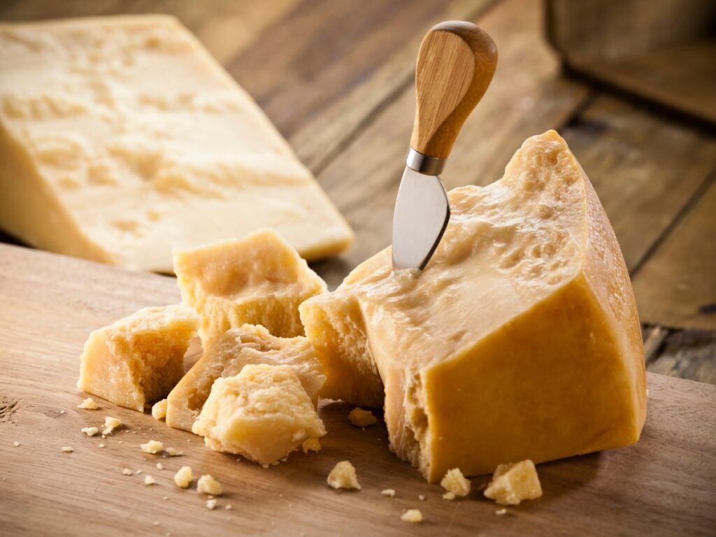 A large wedge of hard aged cheese sits on a wooden board with several broken chunks and a small cheese knife. The firm cheese has a pale golden color and a crumbly texture.