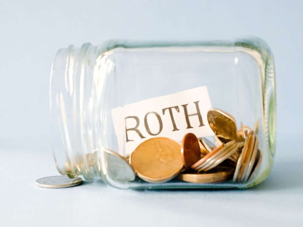 A glass jar on its side is filled with coins and a card reading "ROTH". The savings setup represents money being moved or set aside for a Roth account.