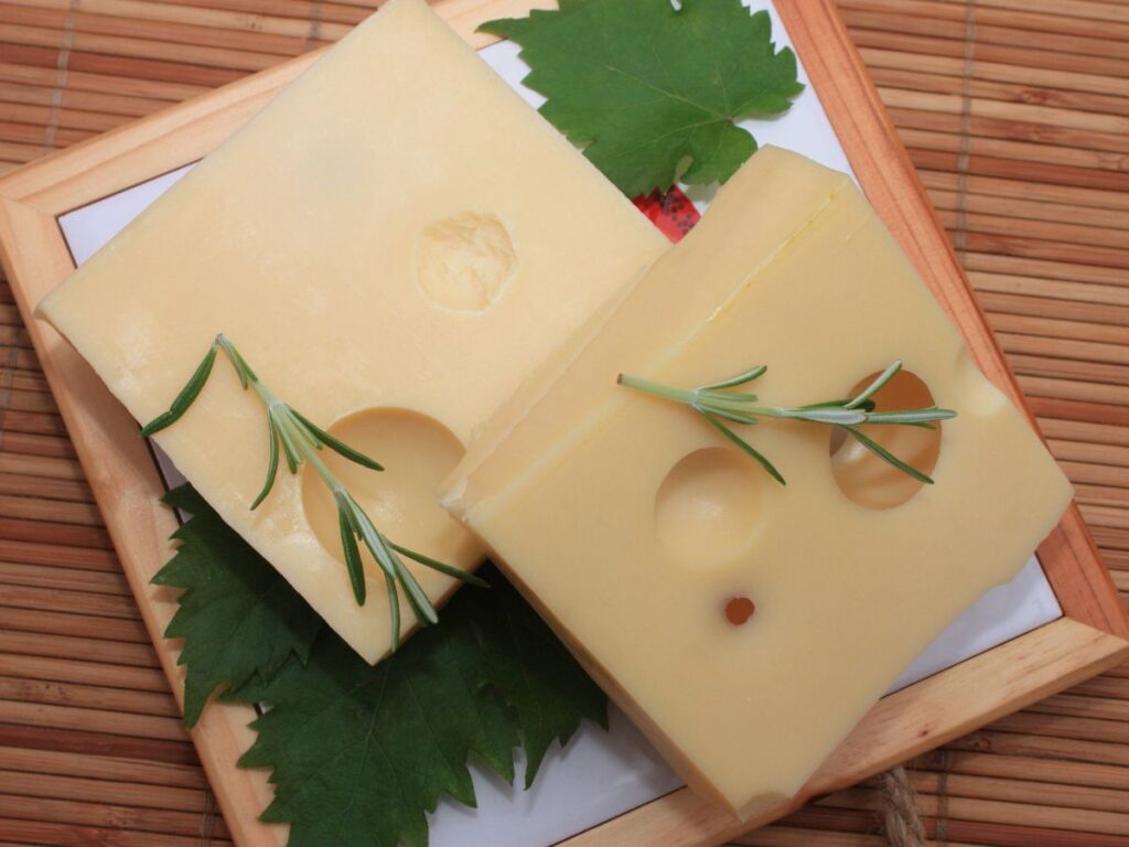 Two thick slices of pale Swiss style cheese with round holes are arranged on green leaves and topped with fresh rosemary. The cheese is served on a small wooden tray.