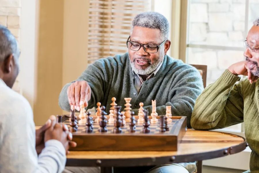 Three older men sit around a table playing chess while one player reaches forward to move a piece. The strategy game represents concentration problem solving and mentally challenging activities.