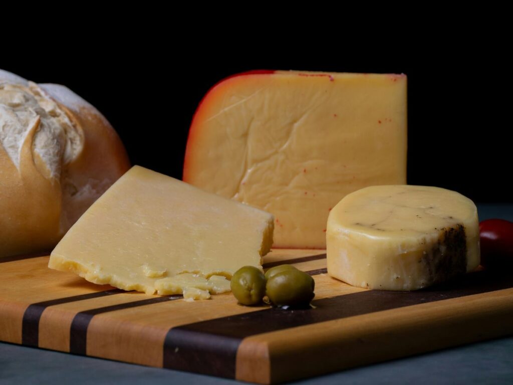 Several varieties of firm cheese are arranged on a striped wooden cutting board with olives and a loaf of bread. The cheeses range from pale yellow to golden orange and include wedges and a small round.