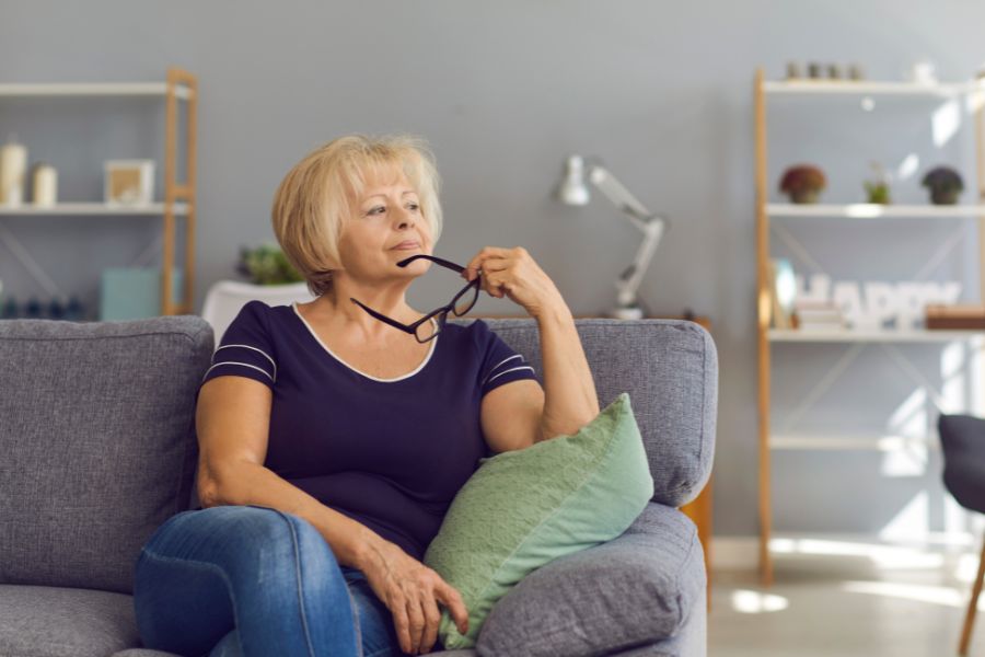 An older woman sits thoughtfully on a gray sofa while holding her glasses near her face. The quiet setting reflects contemplation and maintaining an active mind later in life.
