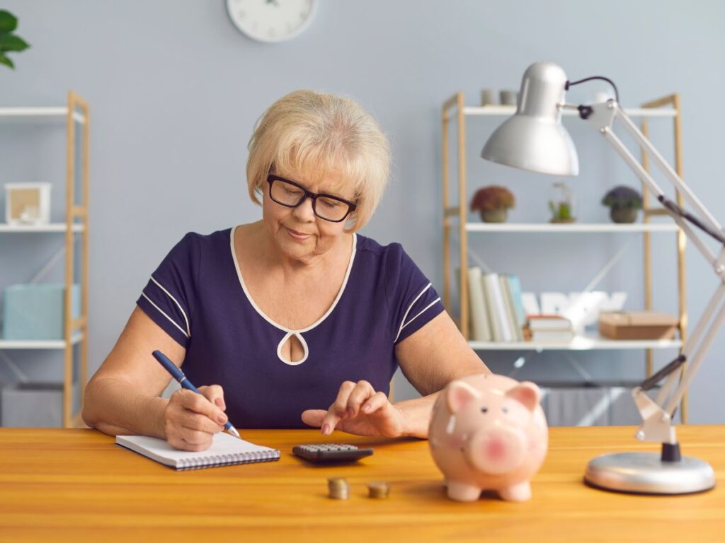 An older woman writes in a notebook beside a calculator coins and a piggy bank. She appears to be reviewing her retirement finances while considering a Roth conversion.