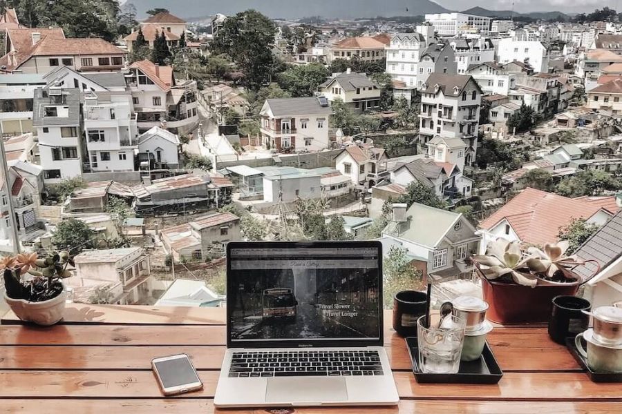 A laptop and phone sit on a wooden table overlooking a hillside town filled with houses and greenery. The laptop screen reads "Travel Slower Travel Longer" and the remote workspace reflects one way people can get paid to travel.