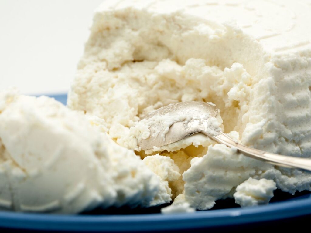 Soft white cheese with a crumbly creamy texture fills a blue plate with a metal spoon resting in the center. This protein rich cheese can be incorporated into meals that support recovery and help rebuild muscle.