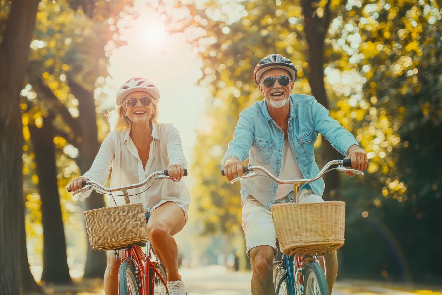 A smiling older couple wearing helmets and sunglasses rides bicycles with wicker baskets along a sunlit tree lined path. The cheerful scene reflects an active outdoor lifestyle for people preparing to retire.