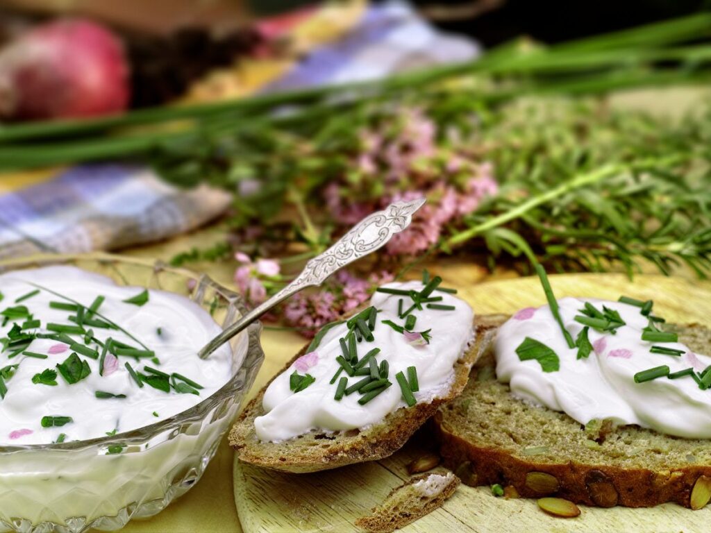 Thick creamy white cheese is spread over slices of dark seeded bread and topped with chopped chives and parsley. More of the herb topped cheese sits in a glass serving bowl beside the bread.