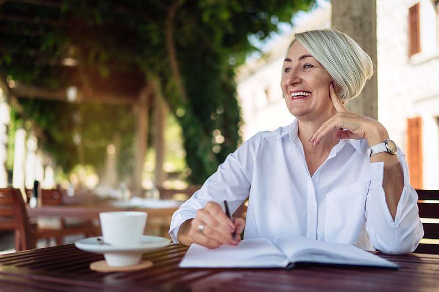 A smiling older woman writes in an open notebook while sitting at an outdoor cafe with a cup of coffee. Writing and continued learning can support the mental engagement associated with How To Raise Your IQ Over 50.