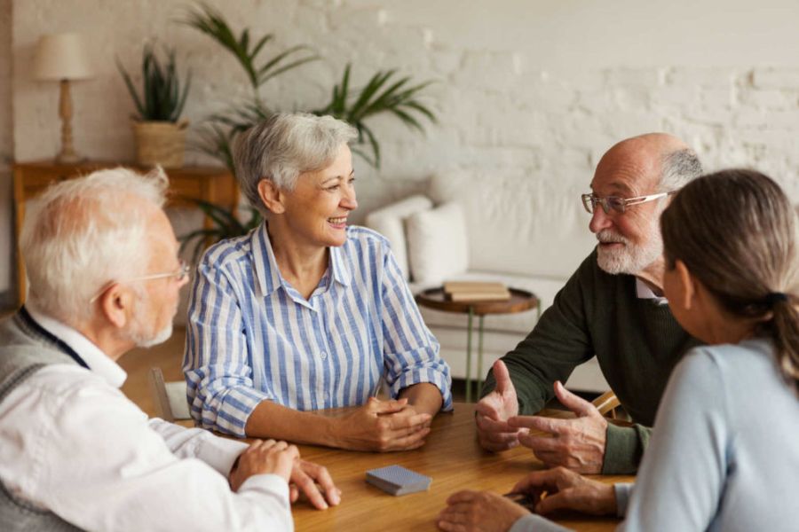 Four older adults sit around a wooden table smiling and having an animated conversation. The social gathering represents discussion connection and mentally engaging interaction.