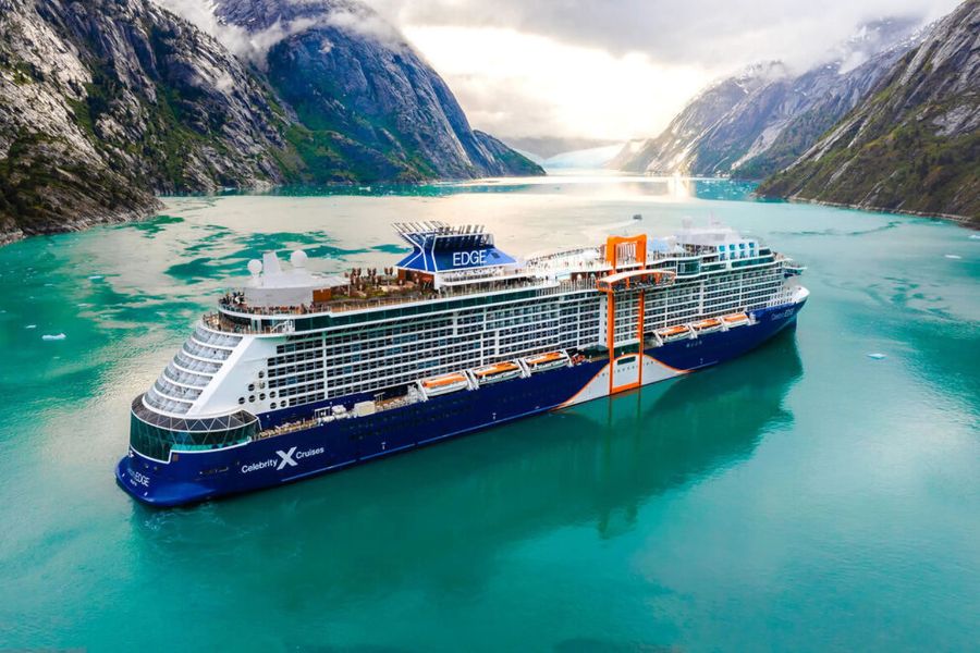 Celebrity Edge sails through vivid turquoise water surrounded by steep rocky mountains and misty peaks. The large modern ship represents the type of voyage travelers may compare when researching cruise discounts for seniors.