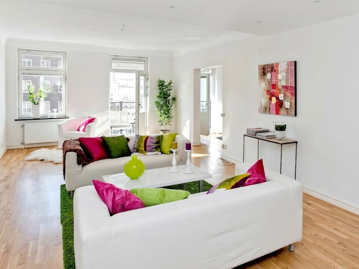 A bright white living room with two sofas arranged around a glossy coffee table and green rug. Pink, green, and patterned throw pillows add strong pops of color against the white walls and light wood floors.