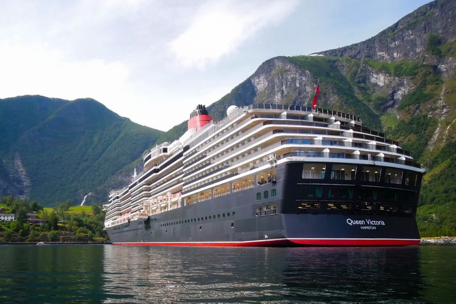 The cruise ship Queen Victoria sits on calm water beneath towering green mountains and rocky cliffs. The stern displays the text "Queen Victoria" and "Hamilton".