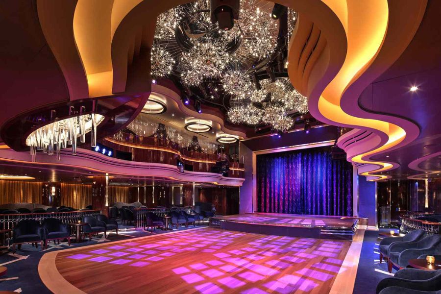 An elegant cruise ship theater features a large wooden dance floor a raised stage and curved balconies. Purple and gold lighting glows beneath dramatic ceiling fixtures and sparkling chandeliers.