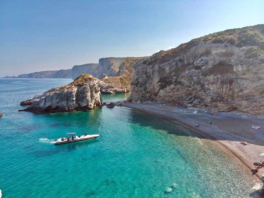 Turquoise water curves around a secluded pebble beach beneath steep rocky cliffs. A small motorboat passes near a large rock formation just offshore.