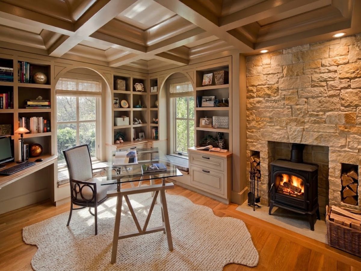A warm home office with a glass desk sits beside a large stone fireplace with a lit wood stove. Built in shelves, arched windows, a coffered ceiling, and natural wood flooring give the workspace a traditional and inviting feel.