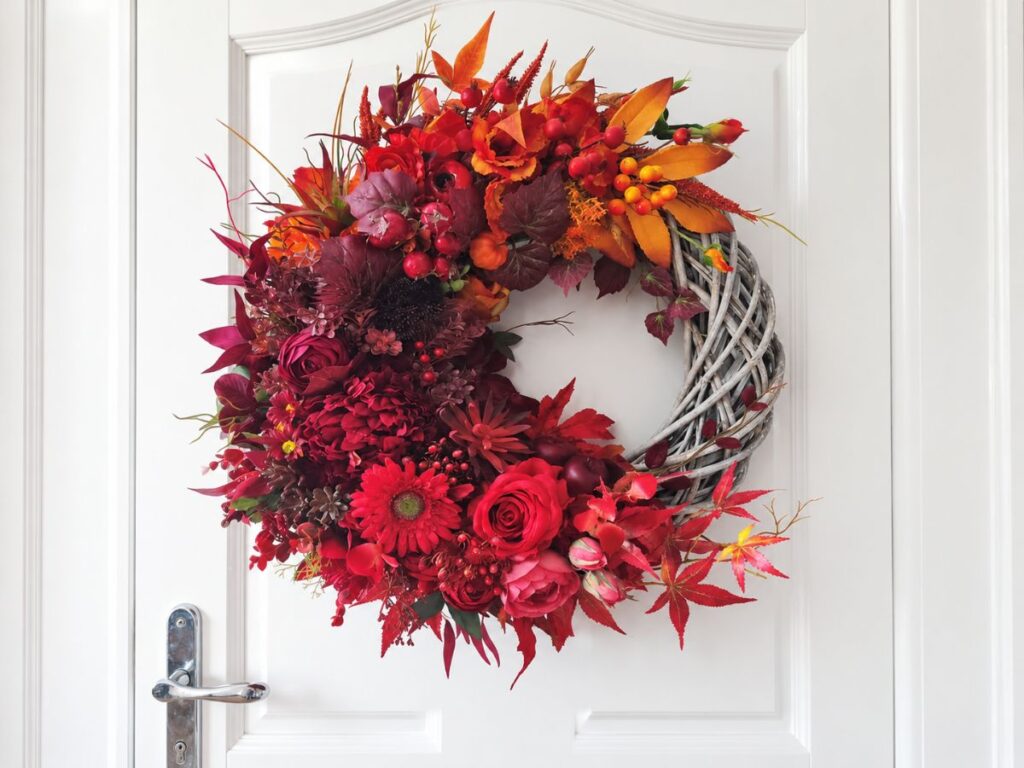 A dramatic floral wreath of deep red roses. dahlias. berries. burgundy foliage. and orange leaves decorates a white front door. Part of the gray woven wreath base remains exposed on the right side for an asymmetrical design.