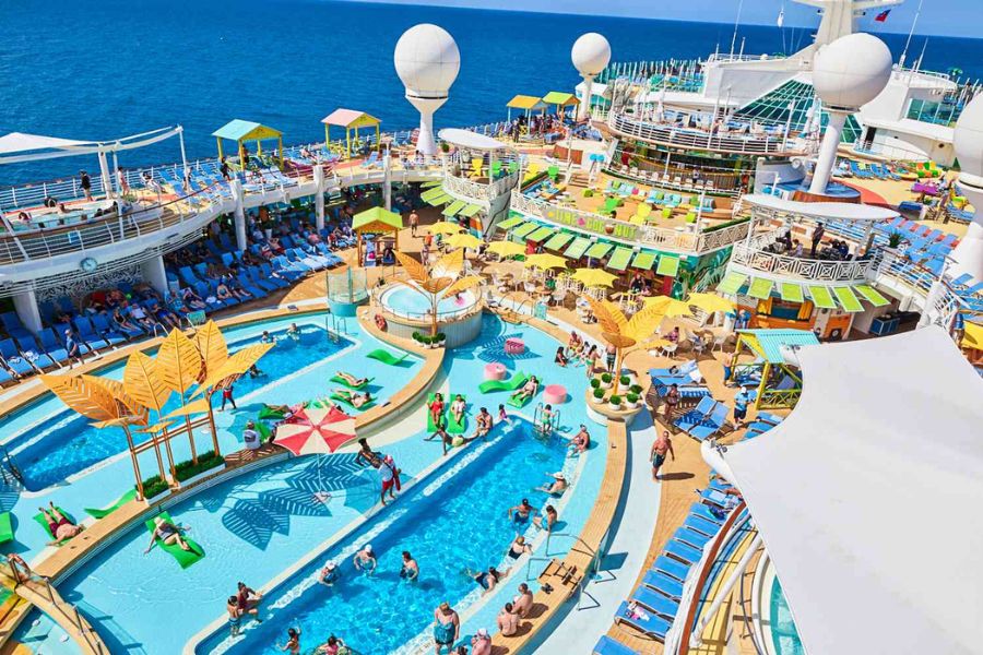 A busy cruise ship pool deck is filled with passengers swimming relaxing on loungers and gathering around colorful shaded areas. Multiple pools bright deck furniture and ocean views show the recreational amenities travelers can enjoy while comparing cruise discounts for seniors.
