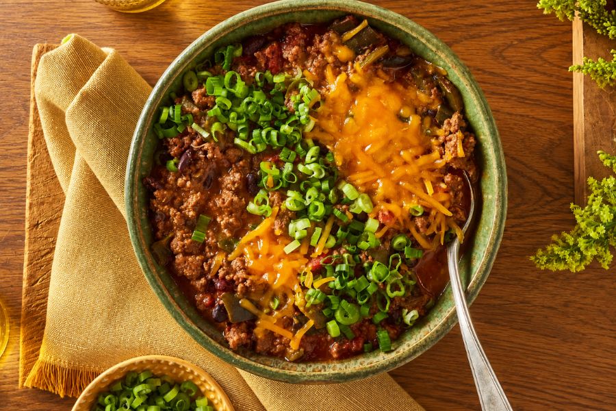 A bowl of hearty beef and bean chili topped with melted cheddar cheese and chopped green onions. The comforting chili is one of the crockpot meals highlighted in the collection.