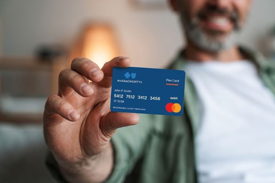 A man holds a blue Massachusetts Flex Card toward the camera while his face is softly blurred in the background. Visible card text includes "MASSACHUSETTS" "Flex Card" "John P Smith" "debit" and "RECHARGEABLE DO NOT THROW AWAY". The card number and expiration details are also visible.