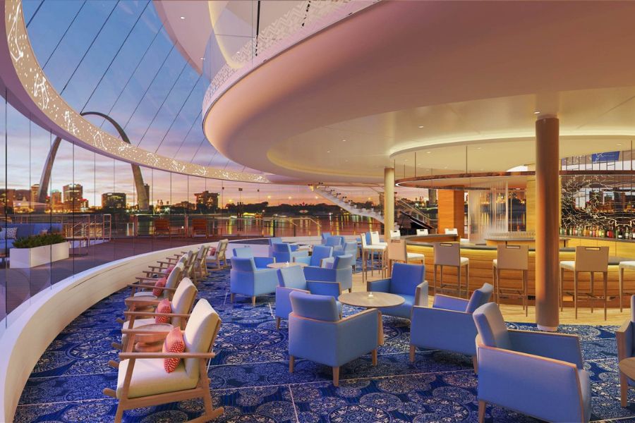 A spacious cruise ship lounge has blue upholstered chairs tables and a curved bar beside floor to ceiling windows. The windows overlook the water and a distant city skyline at dusk.