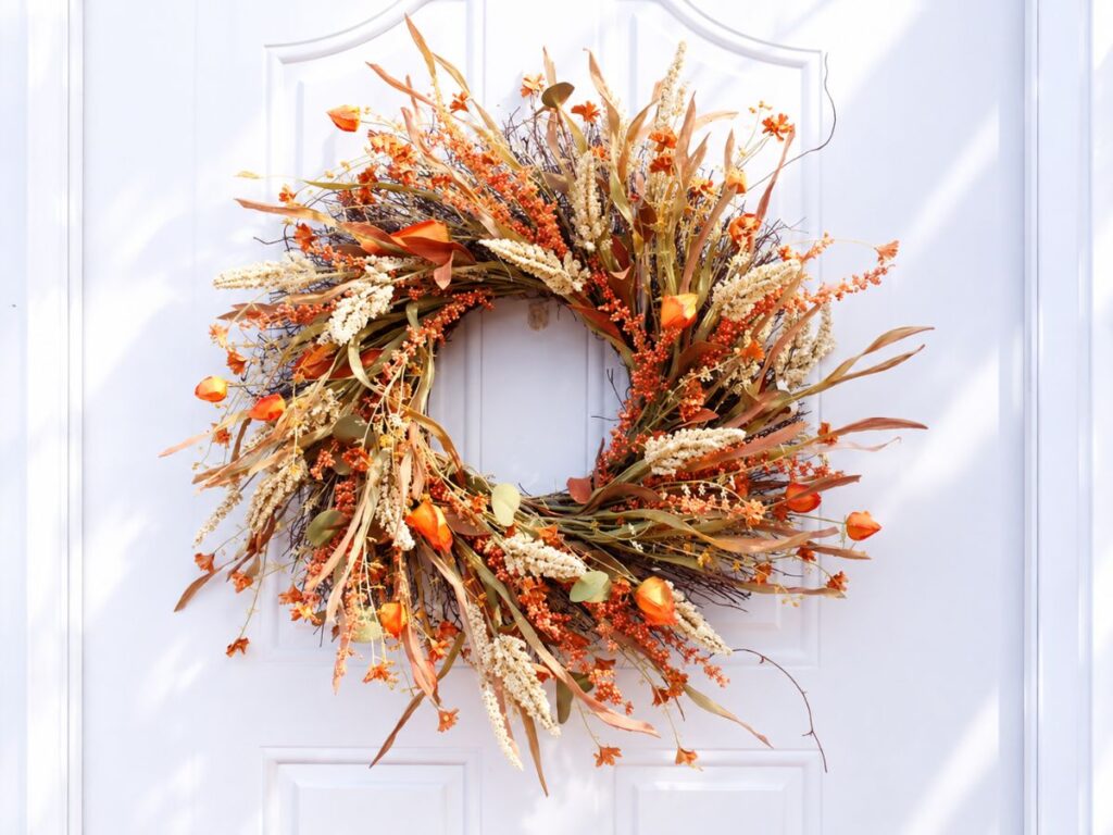 A loose autumn wreath of dried grasses. cream flower spikes. orange berries. small orange flowers. and curled brown leaves hangs on a white front door. This airy example of fall wreaths uses warm seasonal colors and an intentionally untamed shape.
