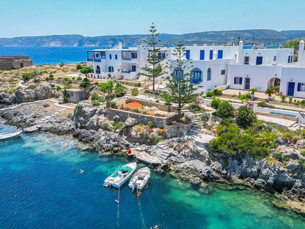 White coastal buildings with blue doors overlook a rocky shoreline and deep blue water. Several small boats are moored in the clear sheltered cove below.