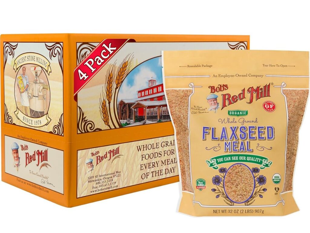A four pack of Bob's Red Mill Organic Whole Ground Flaxseed Meal featuring a beige resealable 32 ounce bag with purple lettering and a clear window showing the ground flaxseed. An orange cardboard display box sits behind the package with illustrations of wheat stalks and the historic mill.