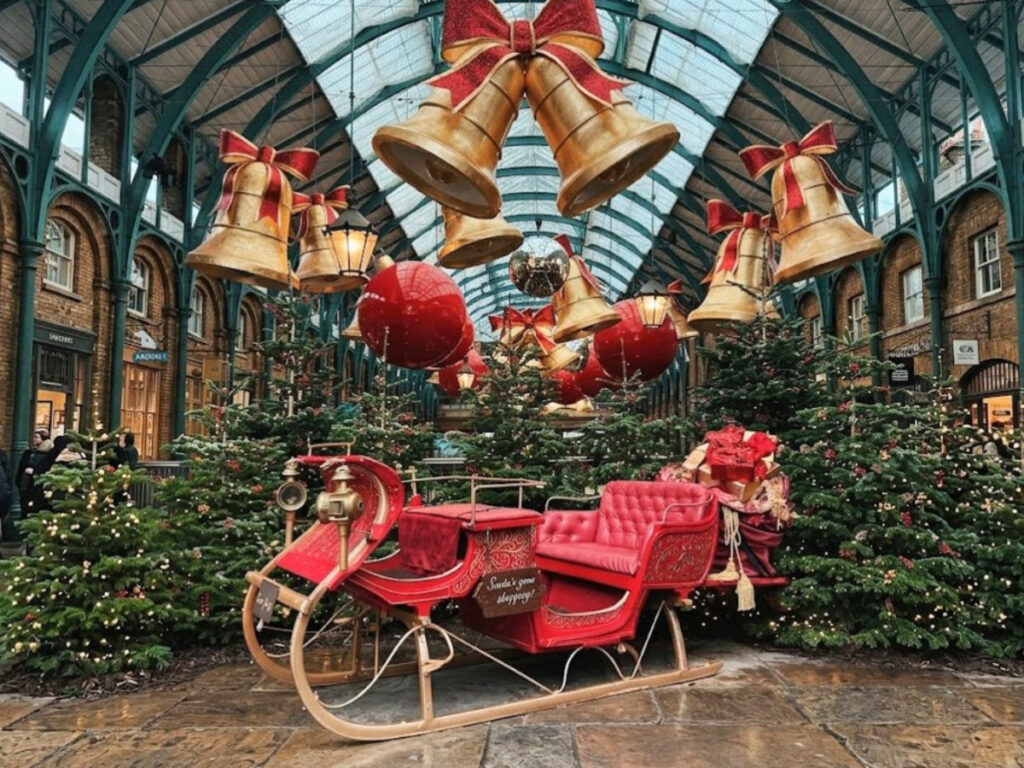 A red and gold Santa sleigh surrounded by decorated Christmas trees inside Covent Garden Market in London. Giant golden bells with red bows and oversized red baubles hang from the glass ceiling as part of the festive Christmas decorations.