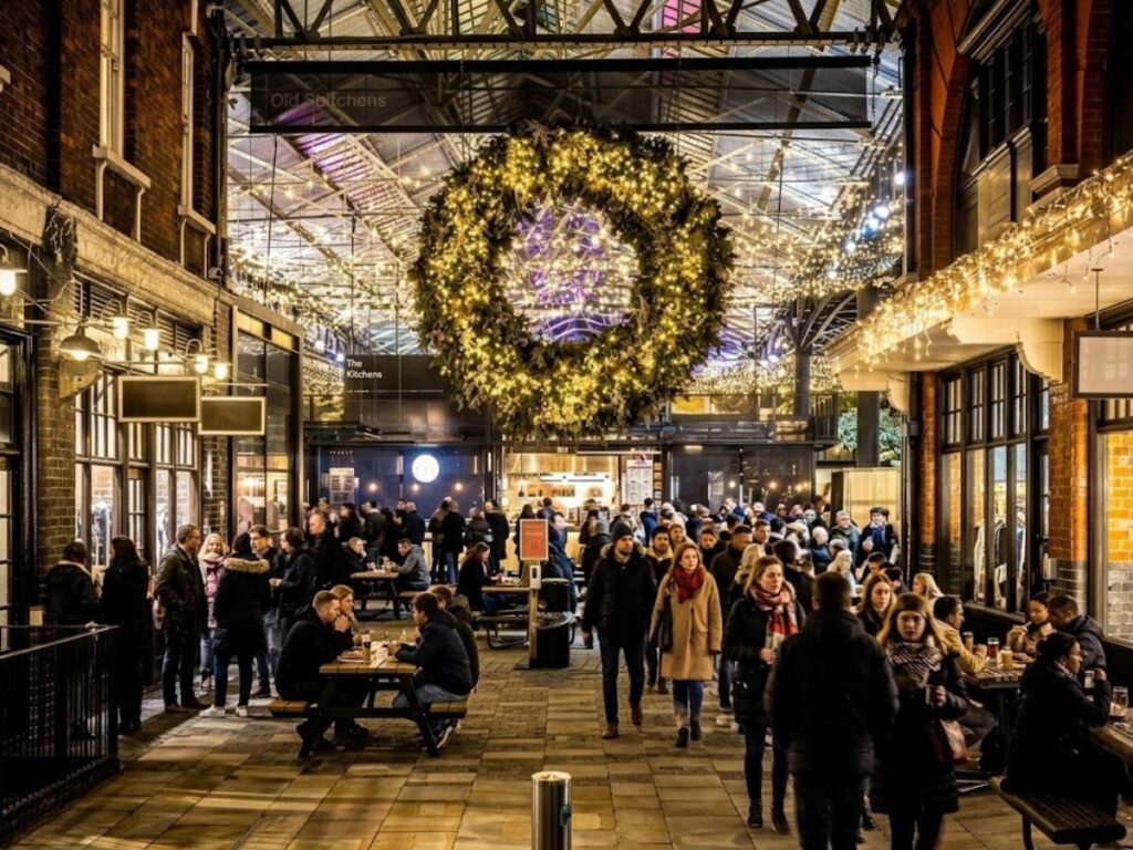 Old Spitalfields Market in London bustling with visitors enjoying food and drinks beneath a giant illuminated Christmas wreath. Warm white fairy lights stretch across the covered market ceiling creating a festive atmosphere during the Christmas season.