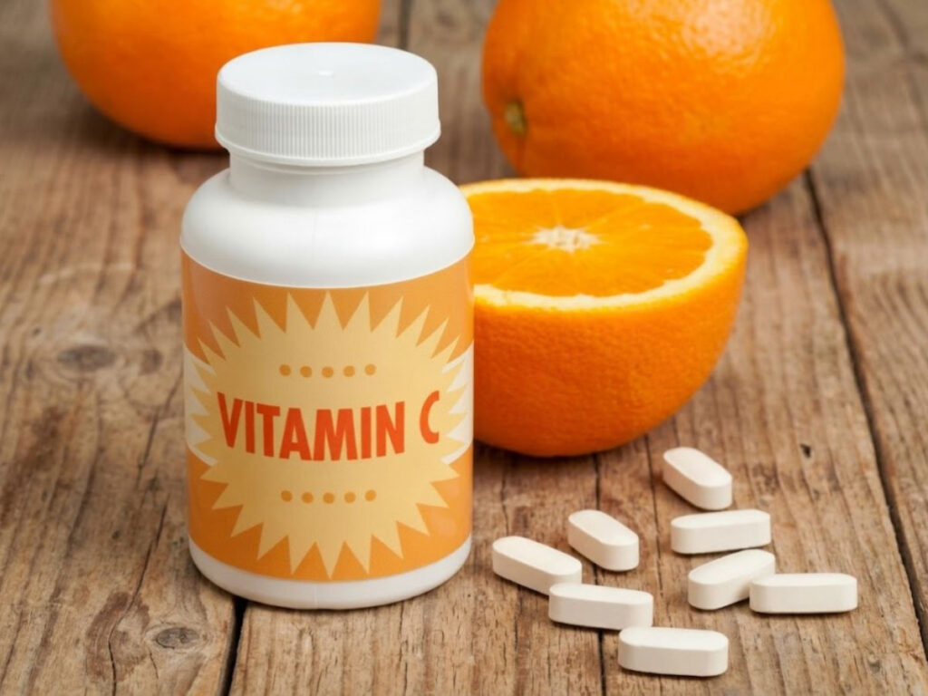 White supplement bottle with an orange label reading "VITAMIN C" sits on a rustic wooden table surrounded by white tablets. Fresh whole oranges and a sliced orange are arranged behind the bottle to illustrate vitamin C supplements alongside a natural dietary source.