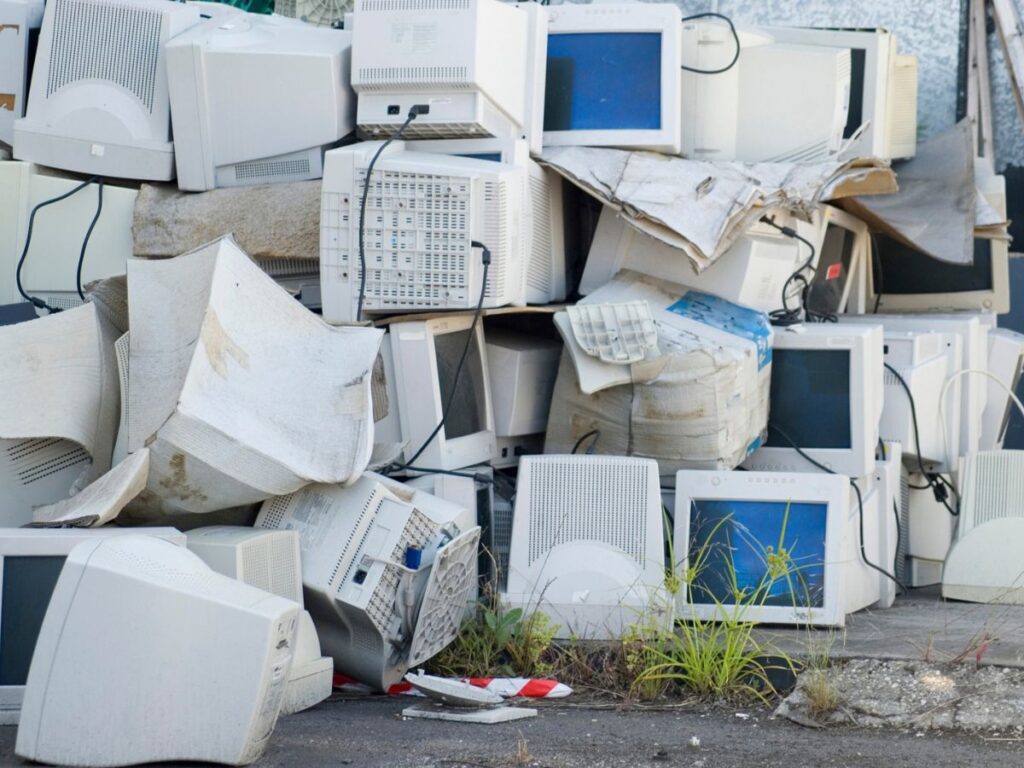 A large pile of e-waste consisting of old computer monitors and bulky electronic equipment stacked outdoors. Broken plastic casings and tangled electrical cables surround the discarded electronics with weeds growing between them.