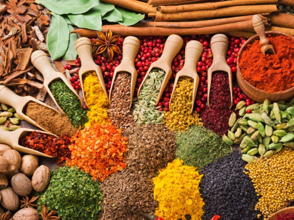 Overhead arrangement of colorful herbs and spices displayed in wooden scoops and scattered across a rustic surface. Cinnamon sticks, star anise, cardamom pods, peppercorns, turmeric, dried herbs and other spices illustrate natural ingredients that can add flavor to meals while supporting a heart healthy diet focused on lowering cholesterol.